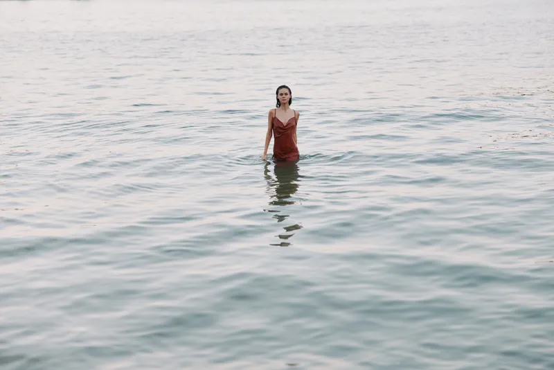 A solitary figure stands waist deep in calm sea water, wearing a vivid red dress that contrasts with blue green tones, creating a serene, cinematic moment at dusk.
