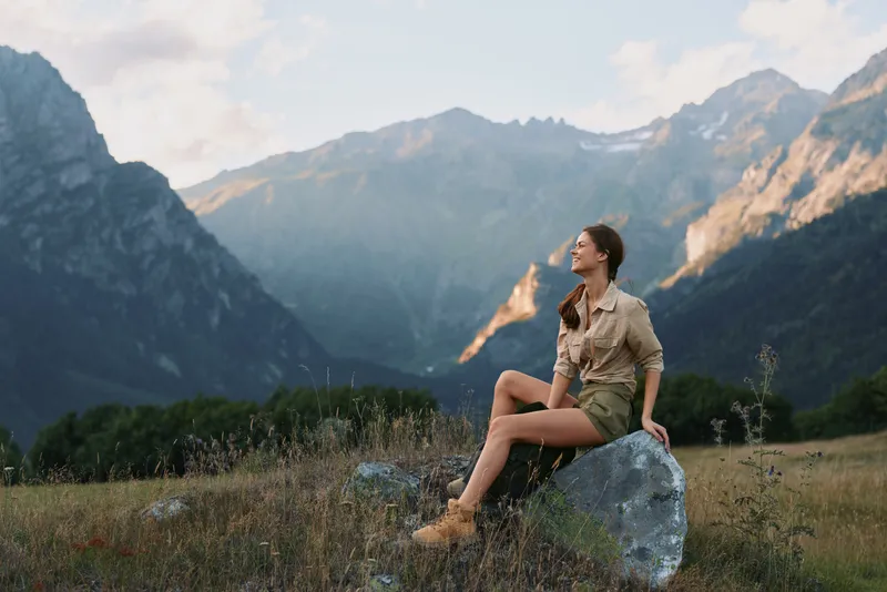 Young woman lifestyle sitting on rock in mountain valley, enjoying nature, relaxed posture, casual clothes, warm sunlight, peaceful outdoor scene, clear sky