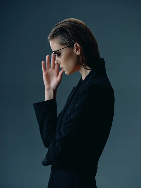 Profile of a thoughtful woman in black suit with glasses on dark background, showing contemplation and style.