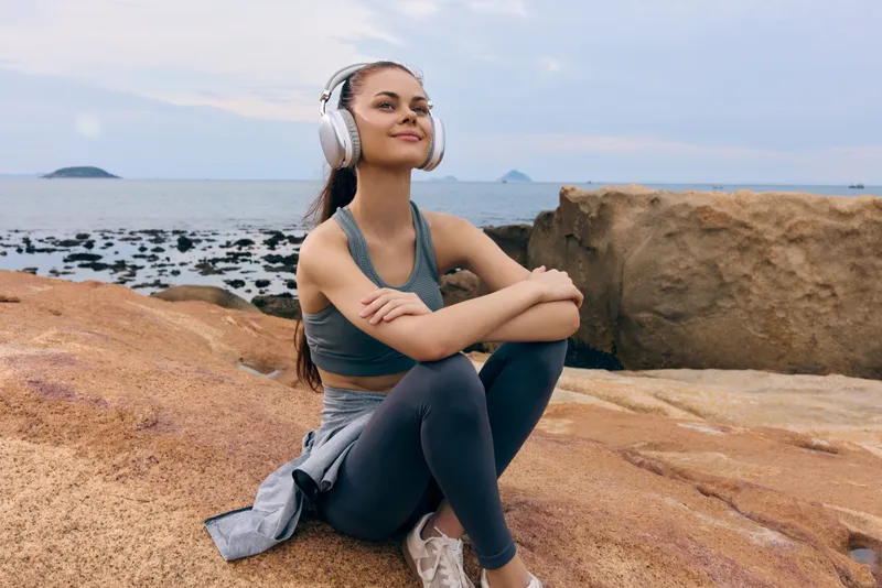 Young happy woman enjoying music outdoors, wearing headphones, sitting on rocks by the sea, sporting a sporty look and a relaxed expression, perfect for wellness themes