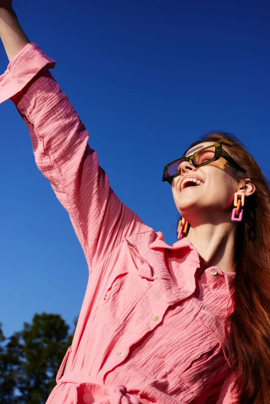 Cheerful young woman in stylish pink outfit enjoying a sunny day outdoors, showcasing vibrant emotions against a clear blue sky, perfect for fashion and lifestyle imagery