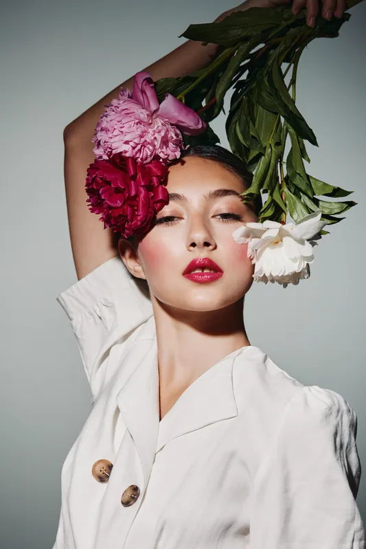 Close-up portrait of a stylish woman with vibrant red lips holding three colorful flowers near her face. Classic beauty and fashion creative concept captured in studio lighting.