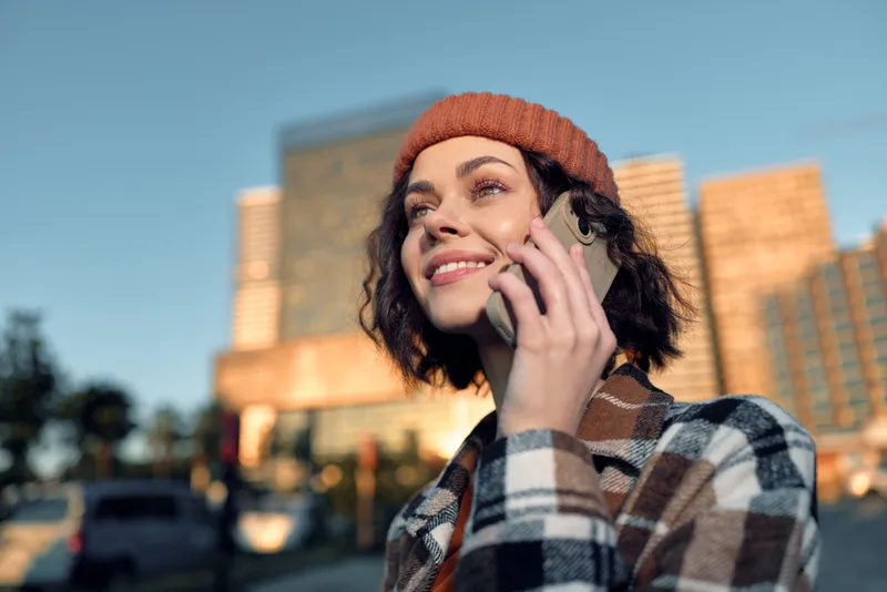Woman on phone with smile in city, wearing beanie and plaid coat at golden hour glow; candid authenticity and mindful living capture emotional storytelling in modern urban lifestyle.