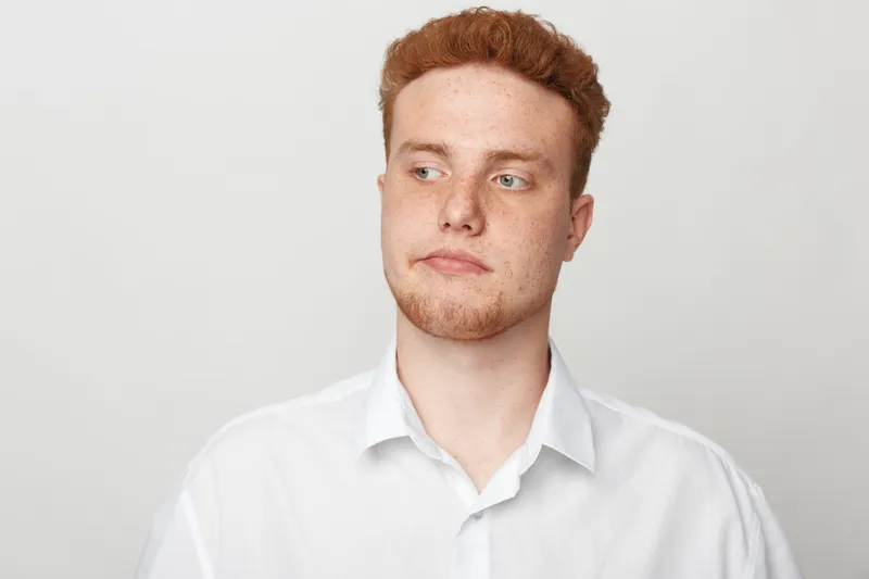 Redhead young man in white shirt looks sideways with thoughtful expression on light background. Concept of contemplation, casual style, and natural portrait of male model.