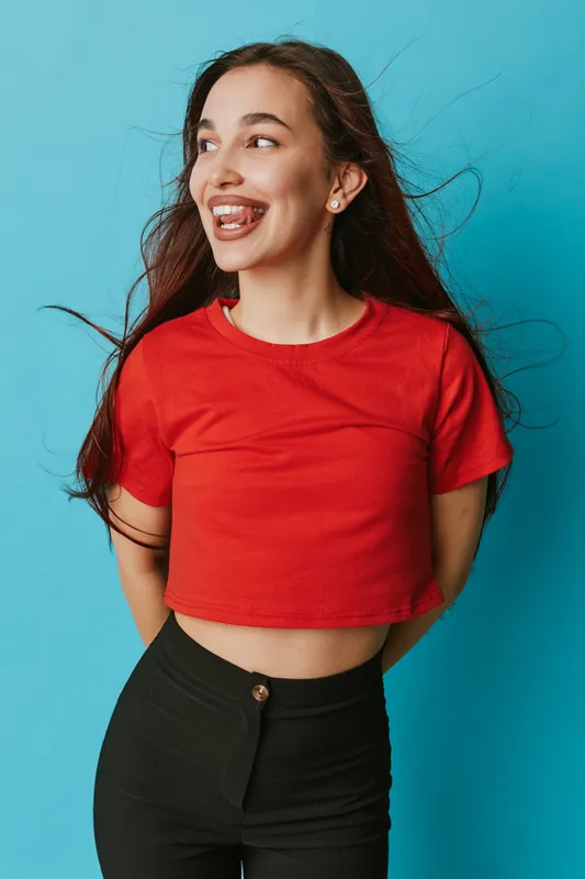 Young woman in red crop top and black pants posing with smile and looking aside on blue background. Casual style, long hair and happy expression with natural light for portrait.