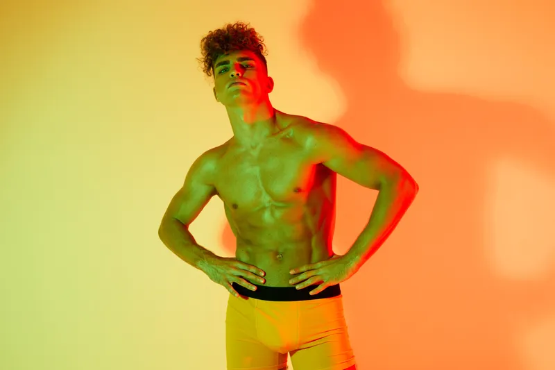 Young muscular man posing shirtless with hands on hips against colorful gradient background, showing confident expression and athletic physique in studio lighting.