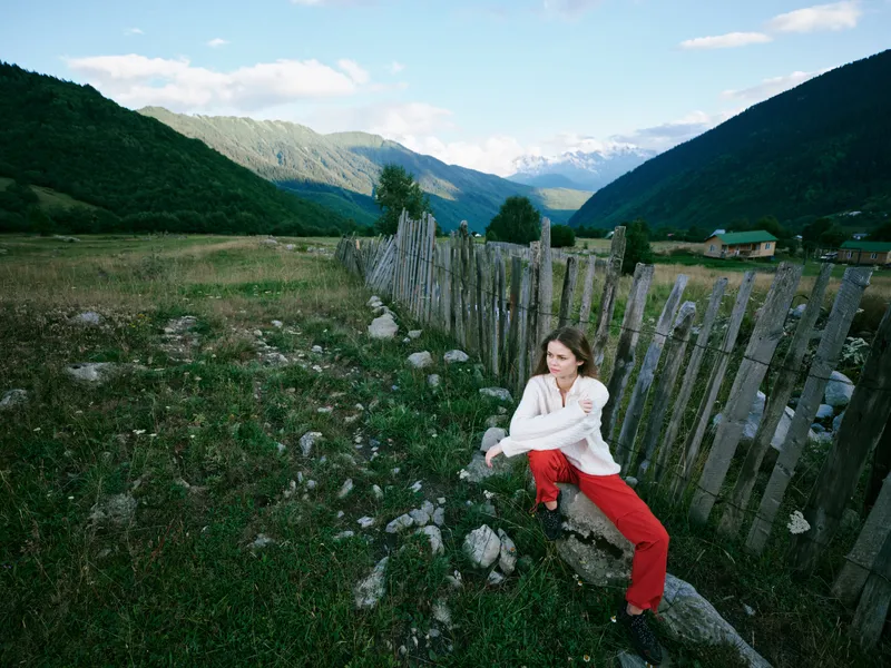 Landscape of rugged countryside with distant mountains, a wooden fence along a rocky meadow, a woman crouching beside stones, white top and red pants, clear sky and tranquil outdoor mood