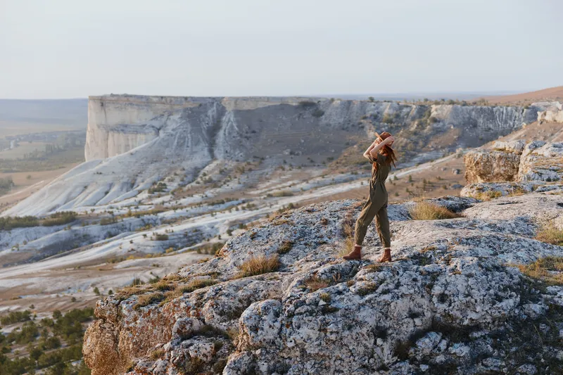 Женщина стоит на краю обрыва, раскинув руки перед собой, наслаждаясь красотой и свободой путешествия