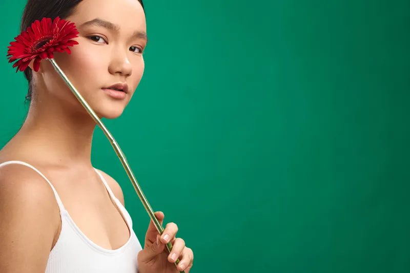 asian woman holds a red flower near her face against a vibrant green backdrop, creating a serene portrait with soft lighting and a calm, thoughtful expression.