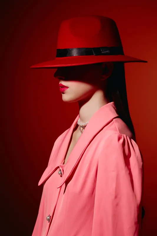 Elegant woman wearing a red wide-brimmed hat and pink tailored jacket stands against red background. Her lips are glossy and makeup is vibrant. Concept of beauty fashion creativity.
