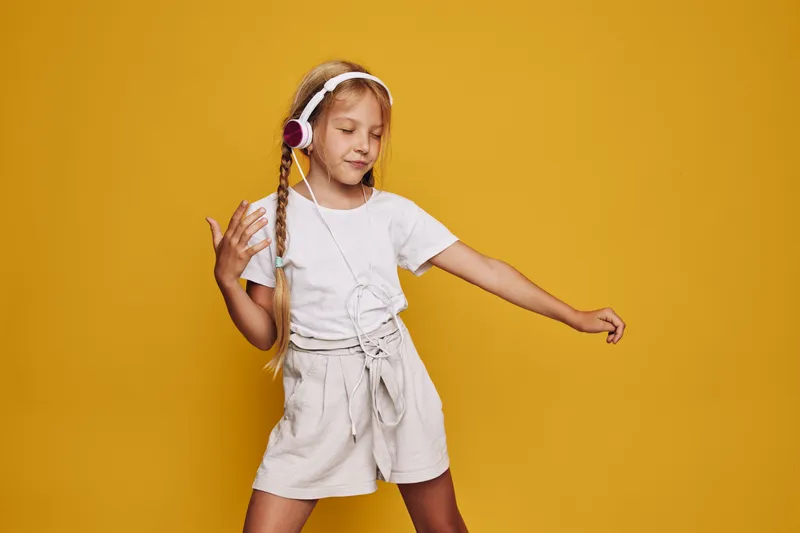 Young girl with braided hair listens to music on headphones and dances joyfully against a solid yellow background. Casual summer outfit.