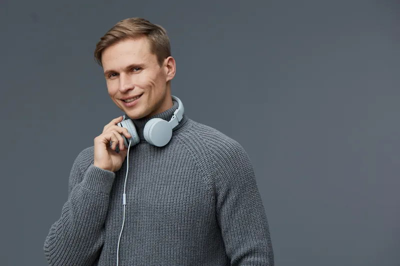Smiling young Caucasian man wearing gray sweater with headphones around neck, holding one headphone near face, looking confident and relaxed, in front of plain gray background, studio portrait. People