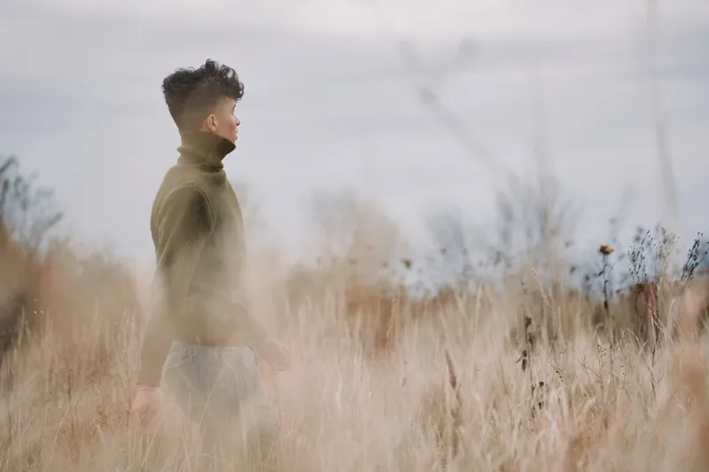 A young man in a turtleneck sweater stands in tall dry grass, captured as a candid, authentic portrait that emphasizes genuine emotion, natural light and credible realistic outdoor detail.