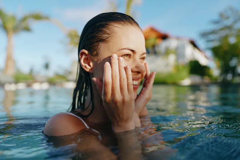 Cheerful woman in a sunlit pool, hands on cheeks, laughing with eyes closed, tropical backdrop, water reflections, summer vibe, relaxed posture, natural colors