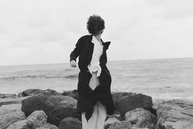 Windy scene of a figure on rocky coastline, wearing a coat and suit as wind tears fabric, black and white mood, ocean backdrop, dramatic movement and fashion oriented coastal editorial
