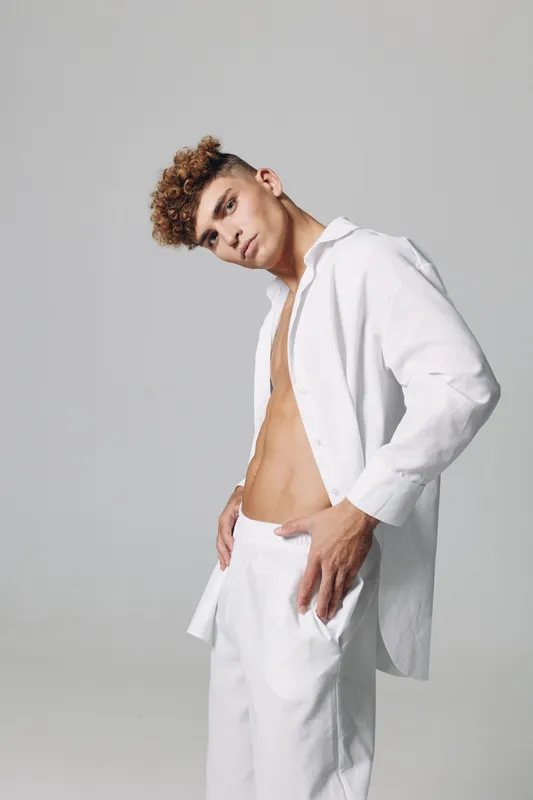 Young man with curly hair wearing white open shirt and pants posing in studio with neutral background. Fashion model showing casual style and confident expression.