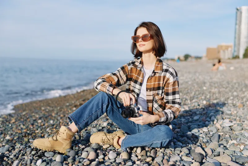 Woman photographer sits on a pebble beach with a camera, wearing a plaid shirt, jeans and boots, sunglasses and a relaxed casual look, exploring light by the sea.