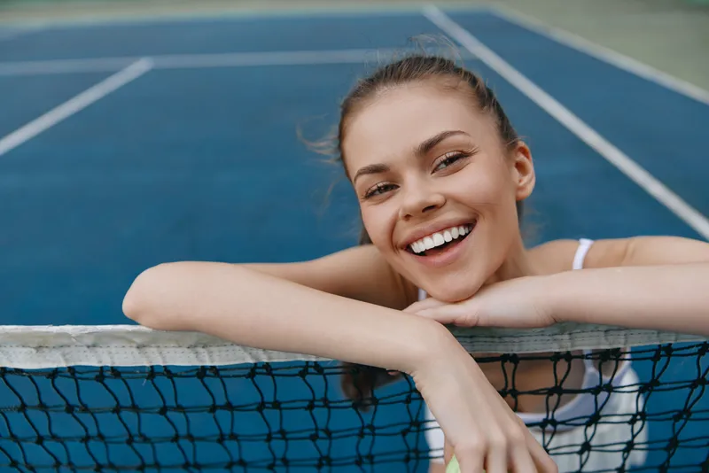 Young female tennis player smiling by the net on a blue court, showcasing joy, sport, and healthy lifestyle for active living