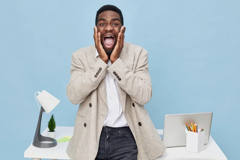 Excited young man expressing surprise with hands on face, standing in modern workspace with laptop, desk lamp, and pen holder, blue background, casual style, positive emotion, indoor environment.