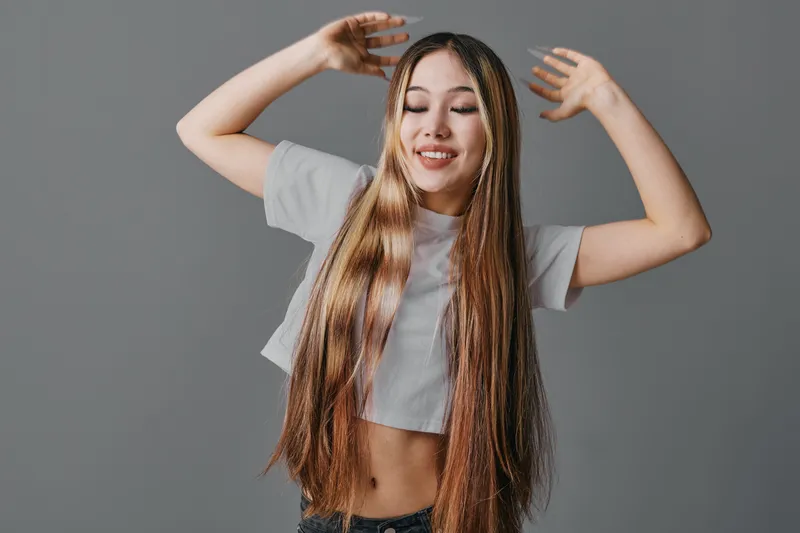 Young woman with long straight hair smiling with eyes closed and raised hands in casual crop top standing against plain gray background in studio portrait.
