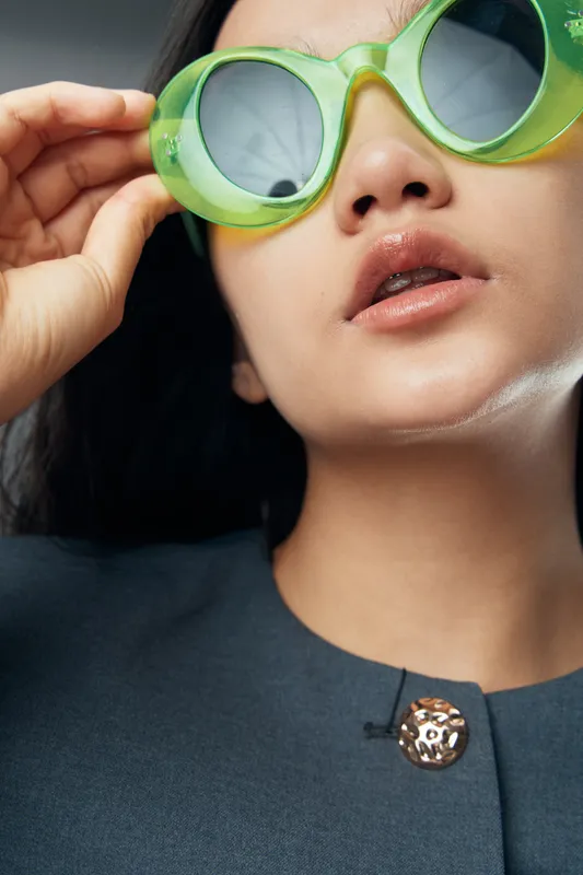 Woman with green sunglasses and glossy lips wearing dark top with decorative button closeup portrait for fashion, style and beauty concept with modern accessories and casual look.