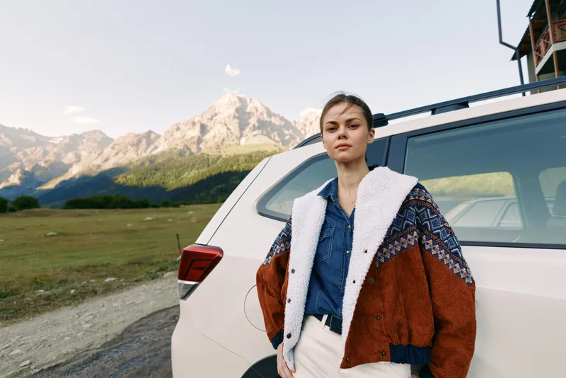 Woman next to car in mountains, travel portrait of young woman in warm jacket leaning against a white SUV on roadside, scenic nature landscape and outdoor adventure.