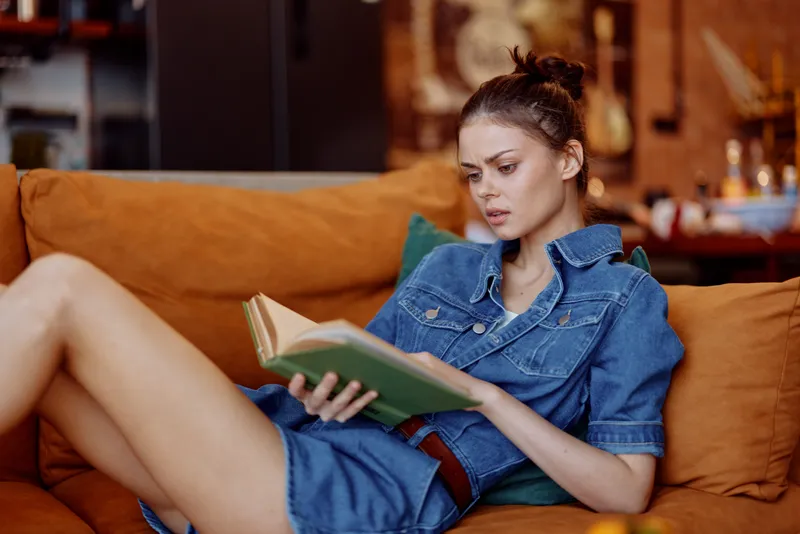 Женщина в джинсовом платье сидит на диване с книгой в руке, читает и отдыхает дома