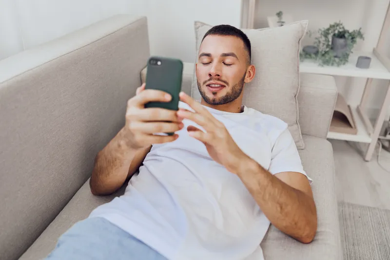 Relaxed man enjoying leisure time while using smartphone at home. Bright living room, casual outfit, contemporary design, and focused expression create a cozy atmosphere.