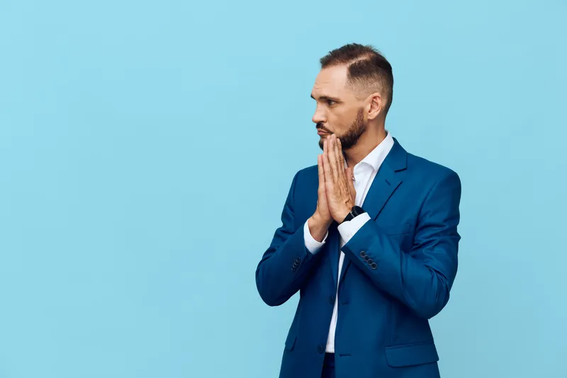 A business professional man stands against a solid blue background, hands clasped near his mouth, conveying deep contemplation, focused intent, and readiness for strategic challenge.