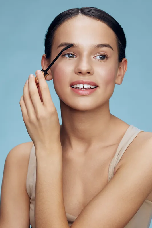 Young woman applying eyebrow gel with a cheerful expression, showcasing beauty and makeup concepts against a soft blue background