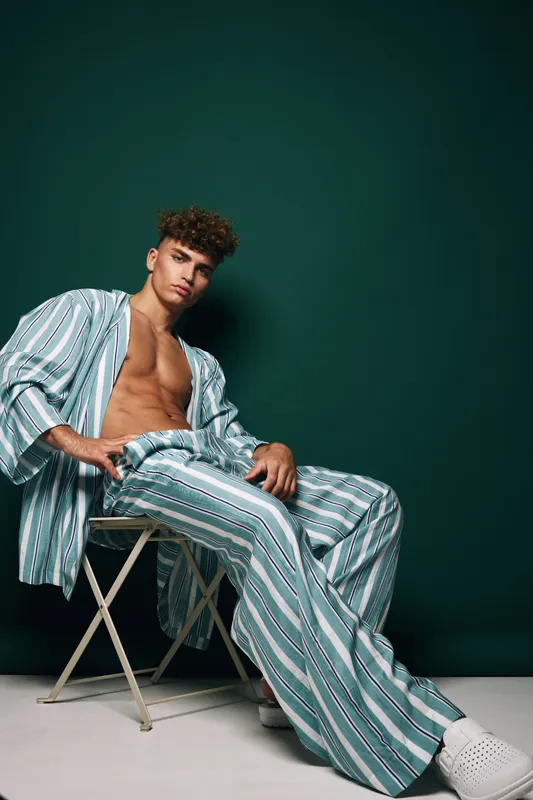 Young man with curly hair wearing striped pajamas and white shoes sitting on a folding chair against dark green background in studio. Fashion, casual, relaxed pose.