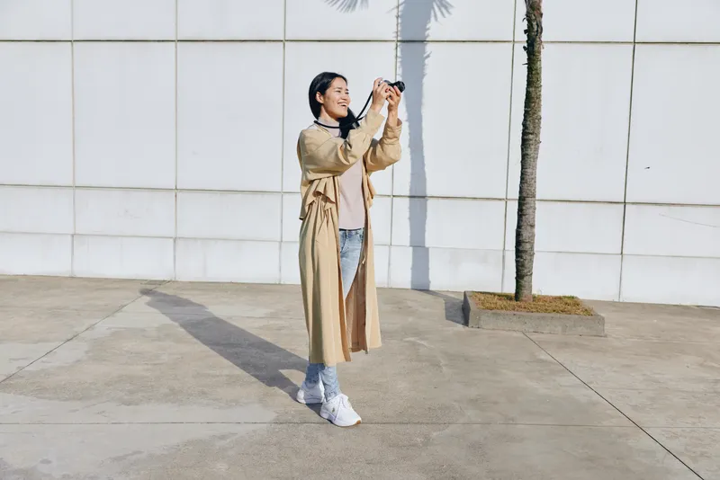 Selfie moment: a woman captures a sunny outdoor scene in a beige coat, denim jeans and white sneakers, standing beside a palm tree against a clean wall, sharing candid fashion and daily life through