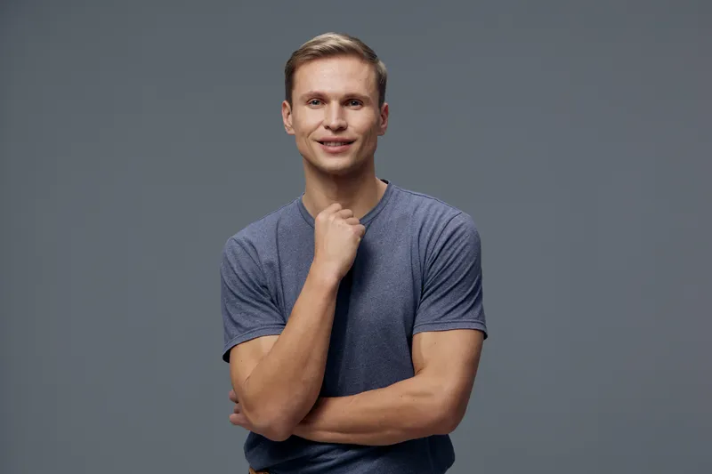 Confident young man in casual blue t-shirt stands against plain gray background smiling and looking at camera, hand on chin, friendly facial expression, lifestyle concept, studio portrait, positive mo