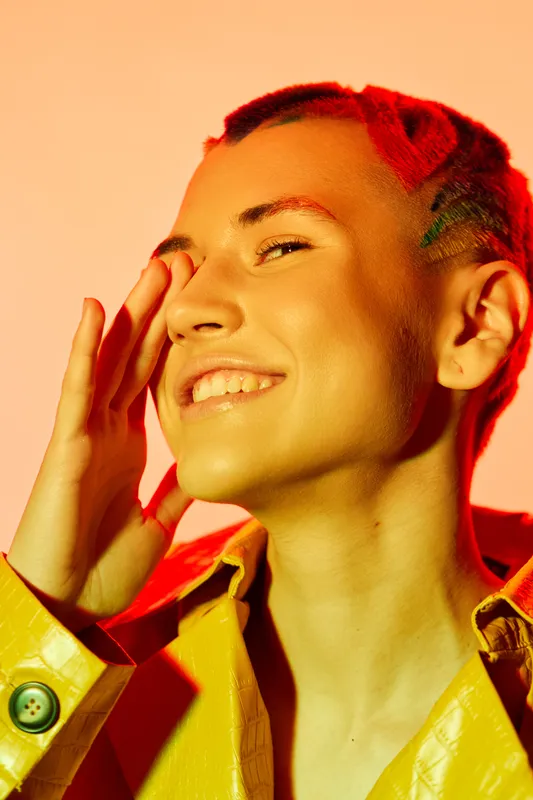 young woman with short hair smiling under warm colored lighting, wearing a yellow textured jacket, expressive joyful mood, studio portrait style
