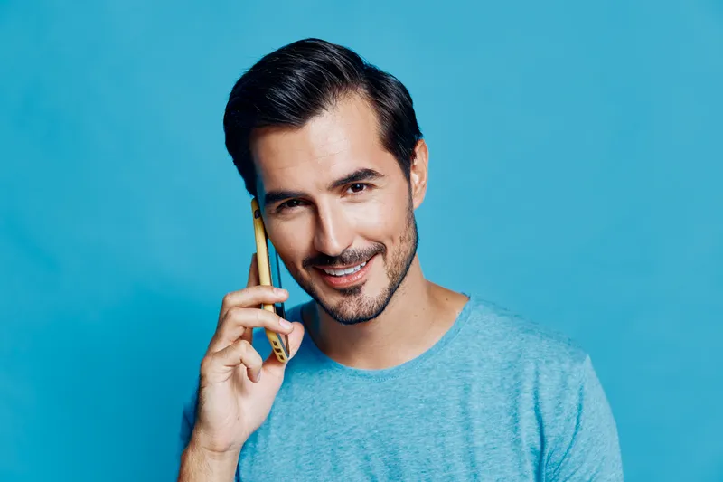 Smiling young man with stylish hair talking on mobile phone, wearing casual blue t-shirt, isolated against a vibrant blue background. Communication lifestyle concept.