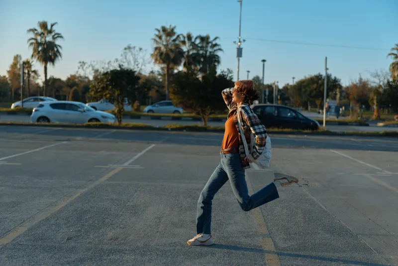 woman running with guitar across parking lot at golden hour, jeans and sweater, backpack swing, sunlight and candid lifestyle mood, authenticity and emotional storytelling vibe