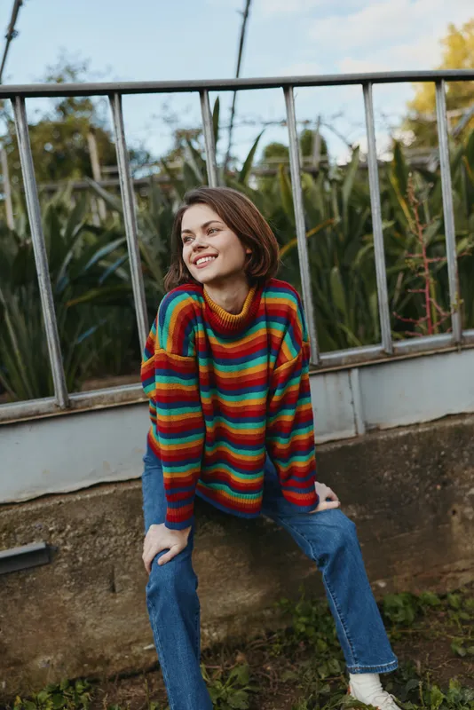 woman smiling in a colorful sweater and jeans sitting outdoors by a metal fence, casual portrait with short hair, relaxed seated pose, natural light and plants in background for lifestyle photography