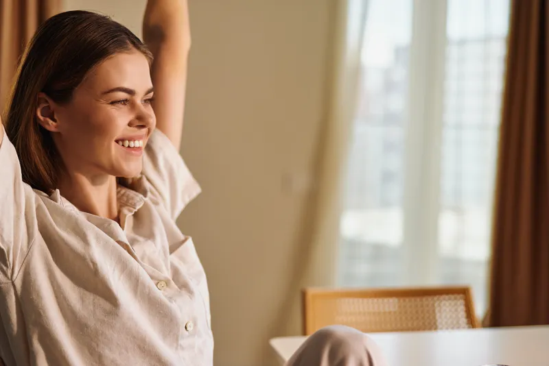 Woman smile stretch at home in morning, relaxed and happy portrait. Casual clothing, natural light by window, cozy interior and comfort for wellbeing and peaceful moment.