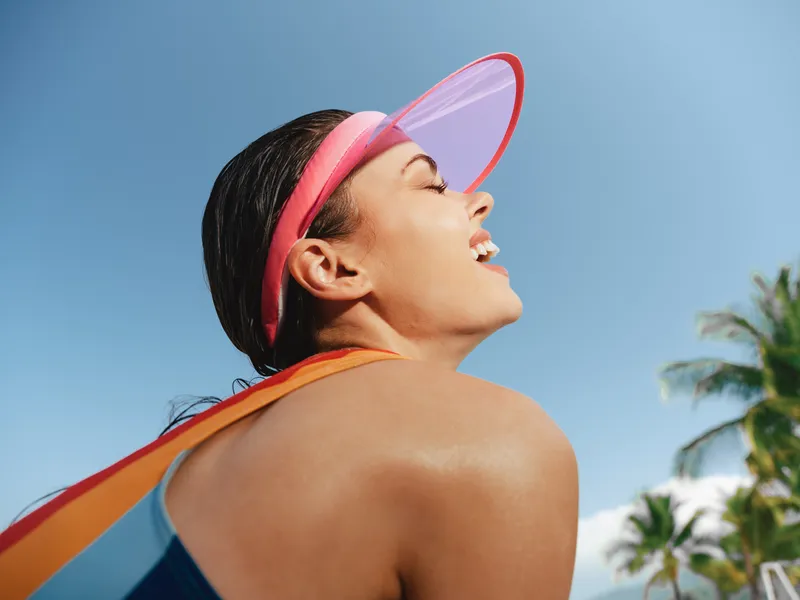 Bright sunlit portrait of a smiling woman wearing a pink visor at a sunny beach, glowing skin, bold colors, and a carefree summer mood.