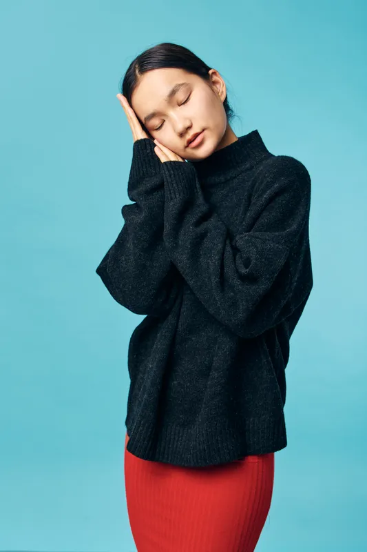 Young woman wearing black sweater and red skirt posing with closed eyes and hands near face against blue background for sleep and relaxation concept.
