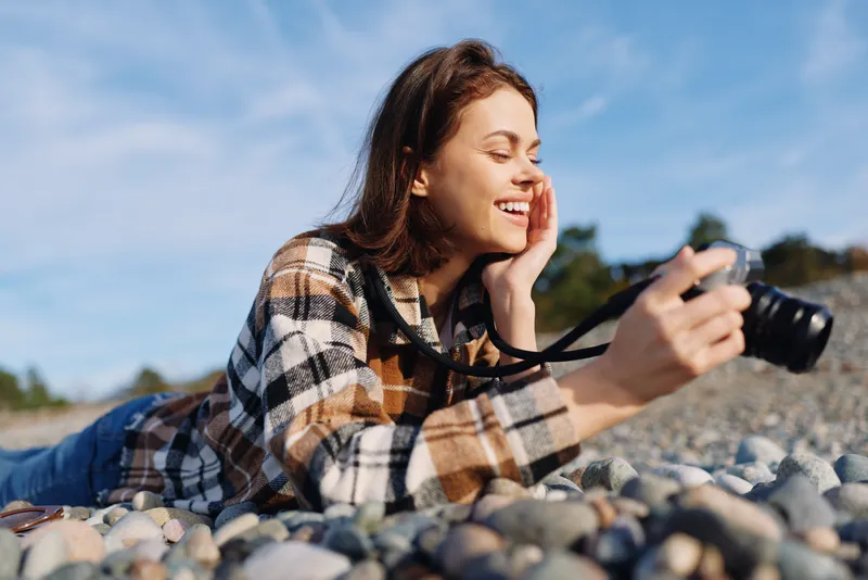 Woman lying on pebbles outdoors, smiling while holding a camera, casual plaid shirt, sunny day, natural light, creative photography moment, relaxed hobby vibe