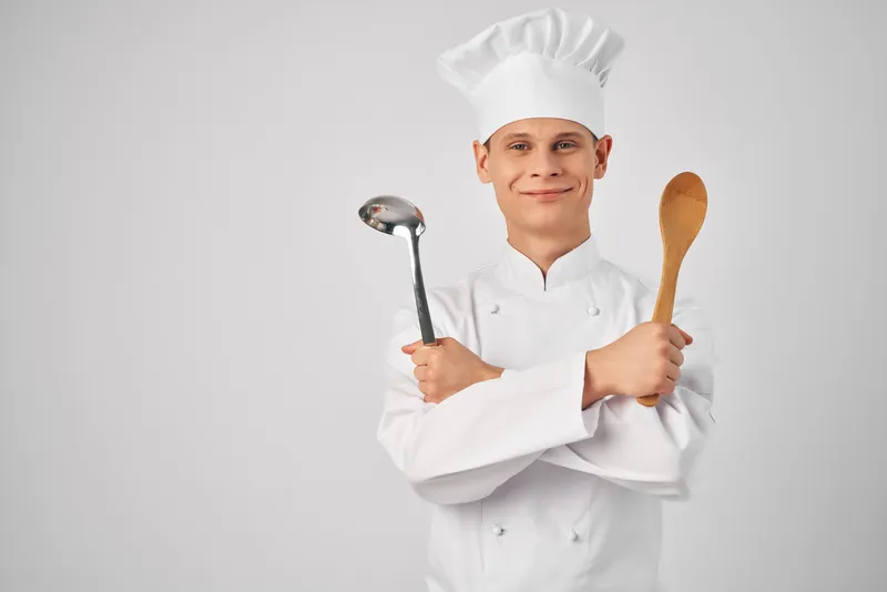 Весёлый шеф-повар мужского пола с кухонной утварью на профессиональном светлом фоне