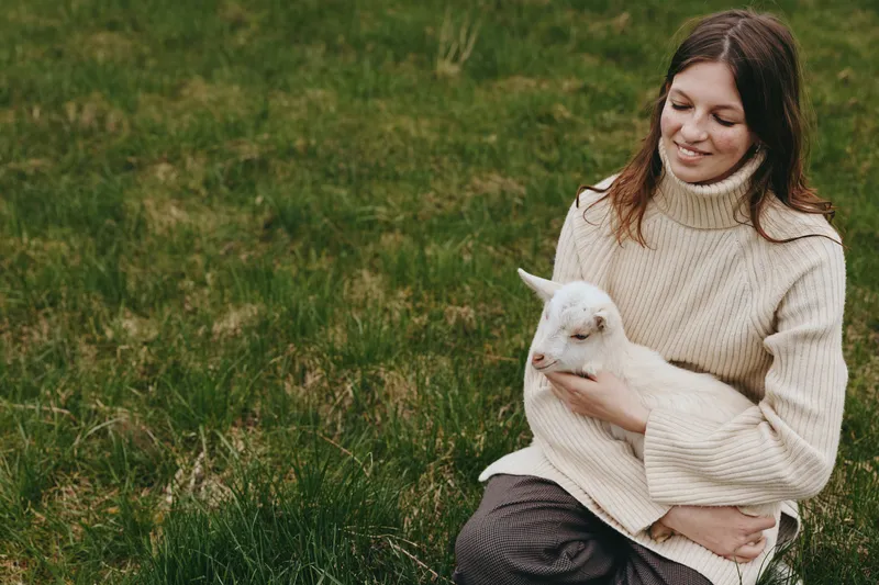 Calm lifestyle scene of a woman holding a goat kid in a grassy field, outdoor moment of affection, warm sweater, natural light, serene connection between human and animal