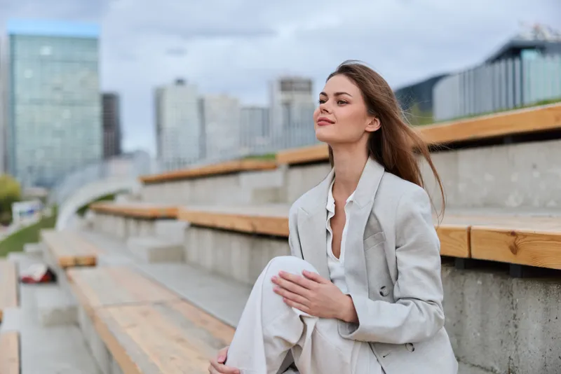 Young woman in light suit sitting confidently on wooden urban steps with modern cityscape background, natural light and relaxed expression