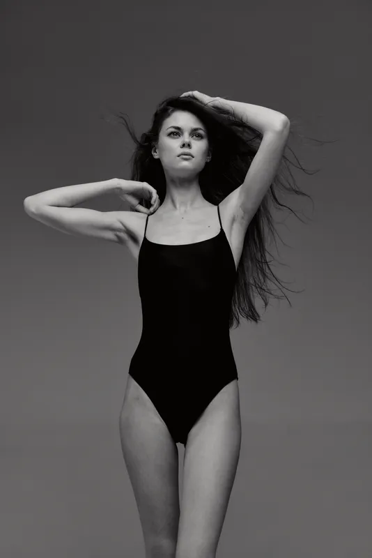 Black and white portrait of a young woman in a black swimsuit posing confidently against a plain background.