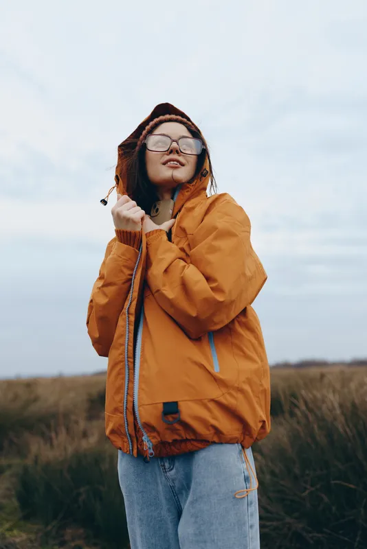 Woman lifestyle outdoors, standing in a grassy field under cloudy sky, wearing orange jacket with hood, glasses and coat for winter cold, relaxed portrait, fresh air mood
