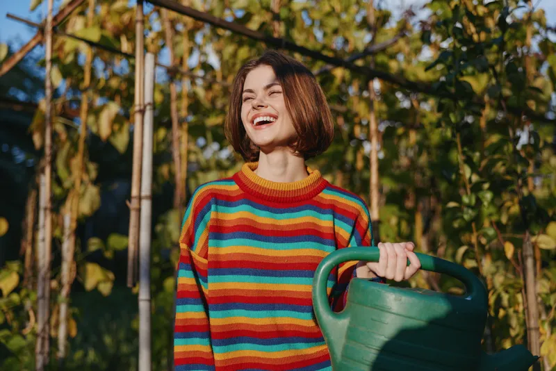Woman smiling with a green watering can in a sunlit garden, young gardener wearing a rainbow sweater and casual outfit, laughing while watering climbing plants and vines.