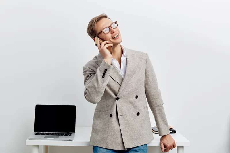 smiling man talking phone casual blazer eyeglasses standing near table laptop modern office digital nomad concept