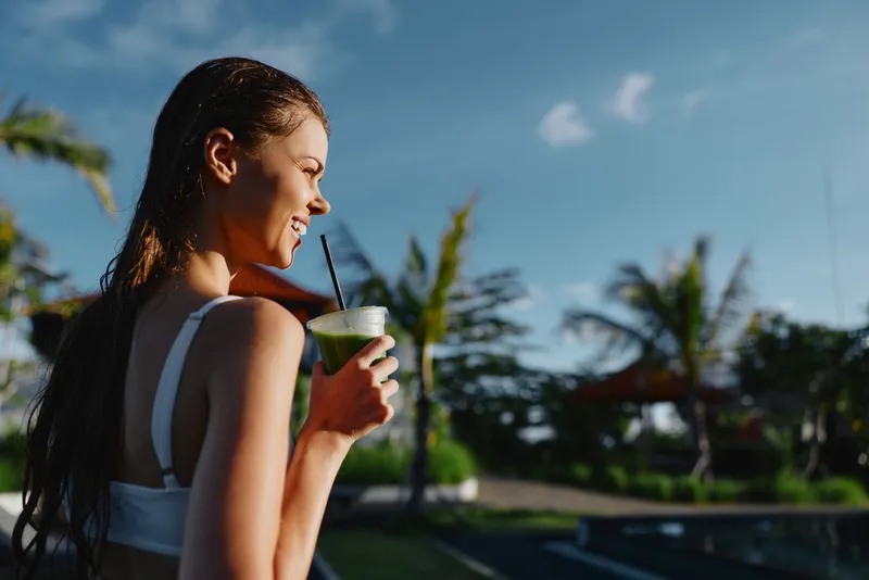 Young woman enjoys healthy green smoothie outdoors in tropical setting with palm trees and blue sky, smiles and holds cup with straw during sunny day lifestyle moment.