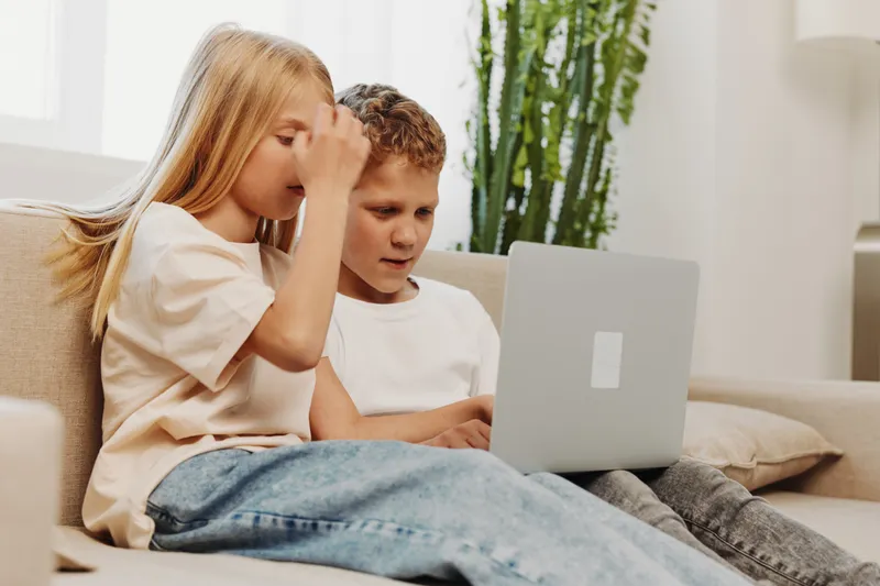 Two focused children sitting on sofa using laptop together in bright home interior with casual clothing and natural light.
